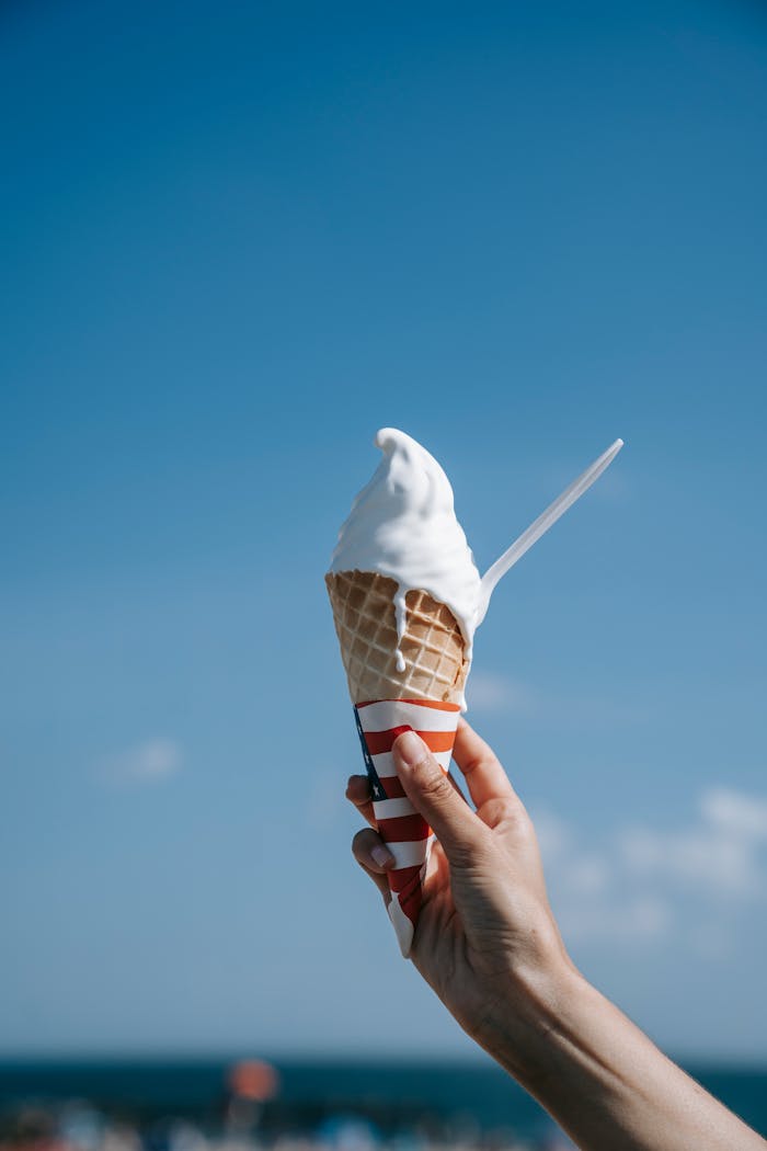 Close-up of a hand holding a soft serve ice cream cone under a clear blue sky.